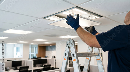 Electrician Standing on Aluminum Ladder Repairing and Installing LED Ceiling Light Panel in Modern Commercial Office Building