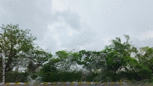 Cinematic slow motion footage of heavy raindrops hitting a car windshield during a rainstorm, with soft green tree silhouettes visible in the background. Captured in ultra high definition 4K at 240fps