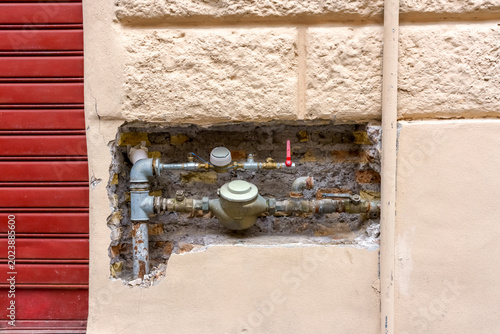 Water meter installation visible on a beige wall with exposed pipes and fittings, red shutter door partially visible in the background