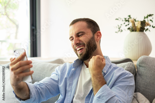 Canvas Print Happy Man Celebrating While Video Calling On Smartphone At Home Couch