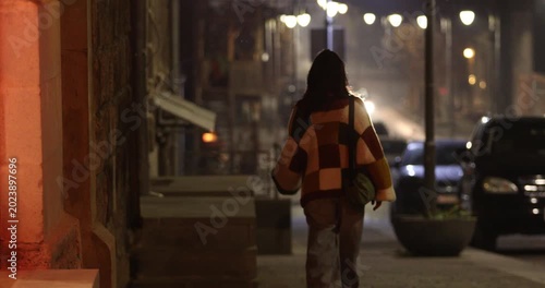 A young woman wearing a checkered sweater walks down a dimly lit, atmospheric city street at night, with bokeh streetlights and parked cars visible in the background, cinematic.