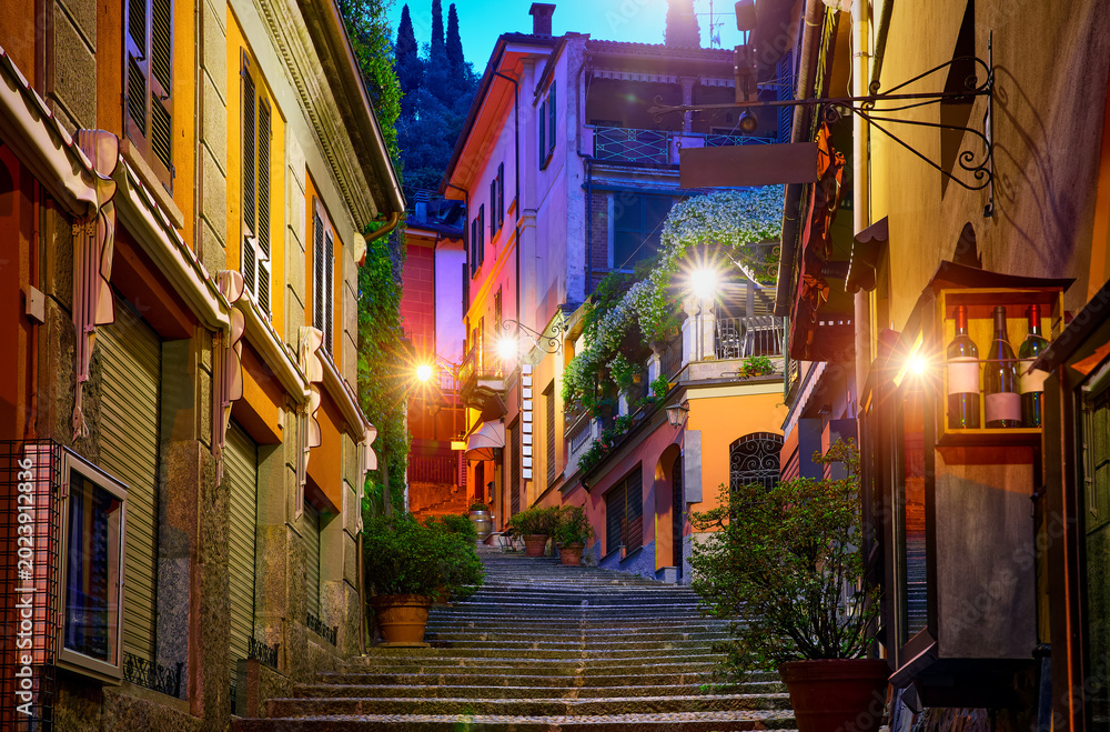 Fototapeta premium Bellagio village at lake Como near Milan Italy, region Lombardy. Night view of famous street with paving stones stairs and cosy restaurants during sunrise with glowing lanterns and old houses walls.