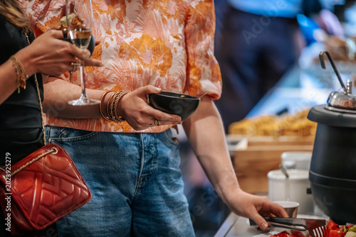 Bilde på lerret Two women at a buffet table, one holding a bowl and a drink, while the other wea