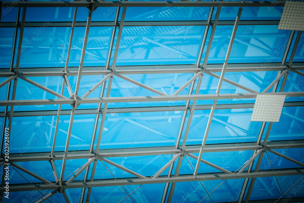 Fototapeta premium Modern architectural ceiling with glass panels and steel framework, showcasing a bright blue sky and natural light filtering through the transparent structure