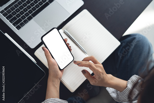 Smartphone white screen mockup, Business woman using smartphone with empty screen with laptop and notebook on table at home office, top view