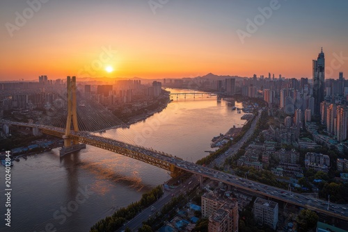 Golden Hour Overlooking a Wide River and Suspension Bridge in a Bustling Cityscape with Traffic and Tall Buildings Under a Warm Sunset Sky