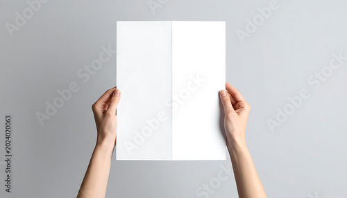 Two hands holding a folded white paper document against a neutral gray background, showcasing a blank template for mockup or presentation