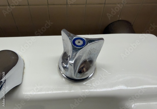 Fotografie Close-up of a dated chrome faucet handle with a blue cap and visible water stains on a white public restroom sink, highlighting concerns about hygiene and maintenance