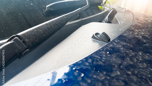 Close-up of a blue car hood covered in water droplets and dust. The image shows the windshield wiper arm and a black washer nozzle. Sunlight reflects on the wet, textured surface of the vehicle.