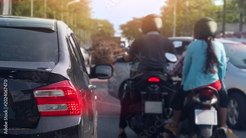 Street scene featuring two people on a motorcycle navigating through slow-moving car traffic. The image captures the mixed mode of transportation commonly found in busy metropolitan areas.
