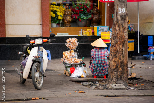 Bild auf Leinwand Street vendor wearing a traditional conical hat selling snacks beside a motorbik