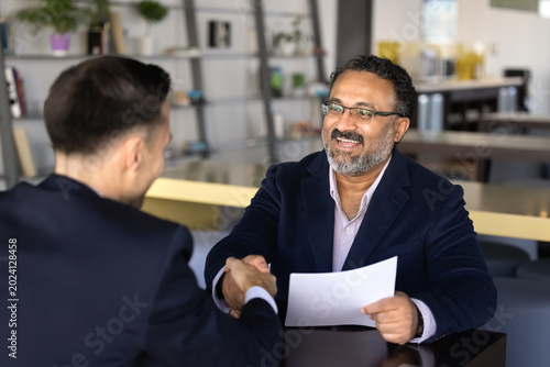 Smiling middle-age hr manager holds resume paper handshakes with male applicant across office table. Business agreement, partnership approval, hiring, commercial deal closure, successful negotiations