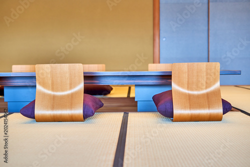 Modern Japanese interior room featuring traditional tatami mat flooring with contemporary curved wooden floor chairs designed for seiza sitting, accompanied by purple cushions. Low blue table