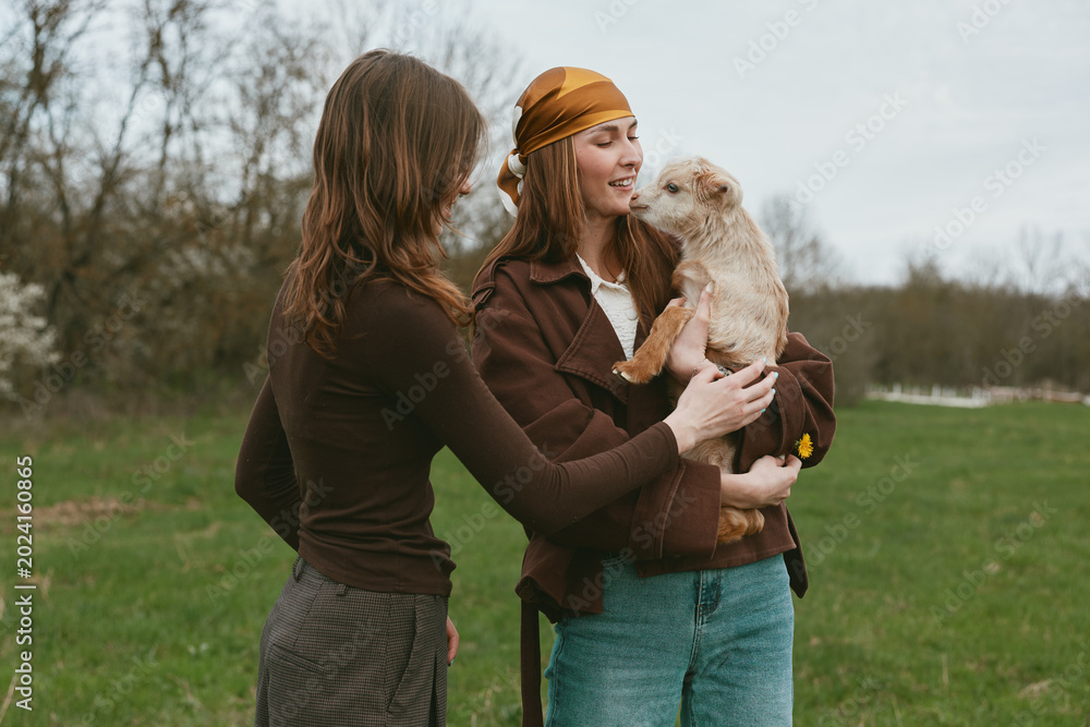 Fototapeta premium Outdoor lifestyle scene with two women smiling as one holds a goat kid, softly lit by natural light, casual jackets, grassy meadow background, carefree connection, warm tones, candid moment.