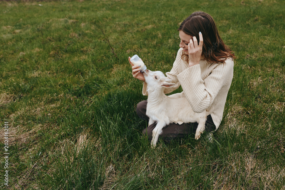 Fototapeta premium A woman sits on grassy ground feeding a young goat kid with a bottle in an outdoor field, capturing a tender lifestyle moment of care, bonding, and simple countryside joy.