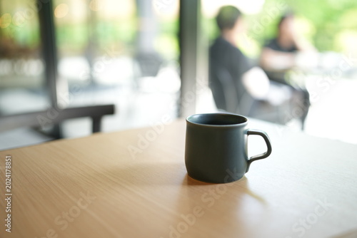 Dark mug rests on wooden table. Soft sunlight filters through glass window. Blurred figures sit in background, relaxed. Quiet, contemplative atmosphere, calm and warm. Ideal for cafe scenes