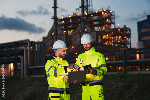 Oil and gas refinery in petroleum industry, industrial plant engineer safety helmet high visibility clothing laptop tablet teamwork night refinery control