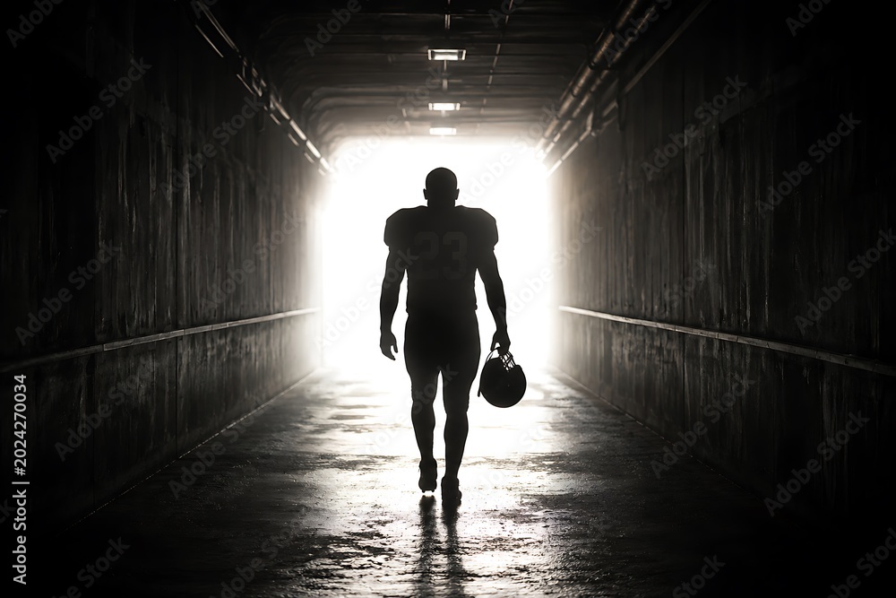 Obraz premium A lone American football player in silhouette walks through a dark tunnel towards a bright light, holding his helmet, symbolizing determination and the path to the field.