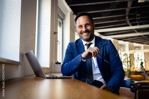 Fotografi Dapper male executive smiling, seated near a window with a laptop, embodying suc