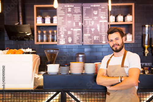 Obraz na płótnie The barista, wearing a white t-shirt and beige apron, has his arms crossed and is confident