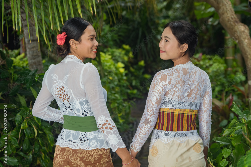 Obraz premium Two Balinese women in white lace kebaya holding hands and smiling at each other in tropical garden
