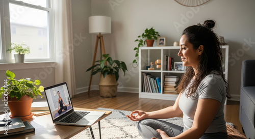 Woman meditating in a bright living room while participating in an online yoga class on her laptop.