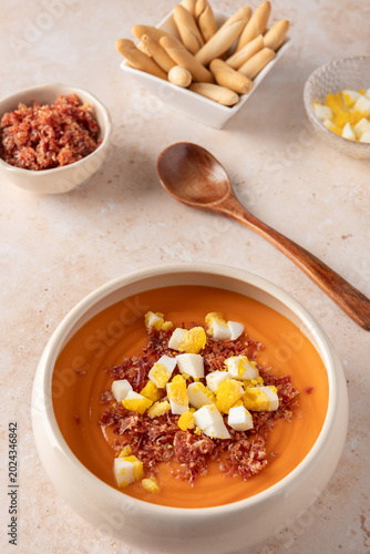 Vertical view of beige bowl with traditional Andalusian salmorejo topped with boiled egg and serrano ham, with ingredient bowls, breadsticks and wooden spoon on light stone background with copy space