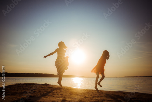 Silhouettes of mother and daughter in dresses jumping and dancing on the beach at sunset. Good relations between generations. Fun family pastime. Health promotion through outdoor activities. Vacation.