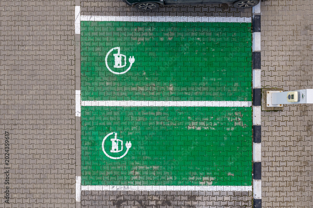 Obraz premium Car charging parking lot. Top-down aerial view of two green eco-friendly electric vehicle charging parking spaces with white painted battery plug symbols on interlocking concrete pavers. Electric cars