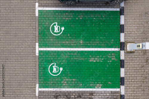 Car charging parking lot. Top-down aerial view of two green eco-friendly electric vehicle charging parking spaces with white painted battery plug symbols on interlocking concrete pavers. Electric cars