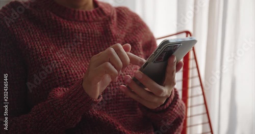 Hand. Asian woman using smartphone on chair by window, scrolling screen with finger in bright home interior, mobile technology and digital lifestyle concept