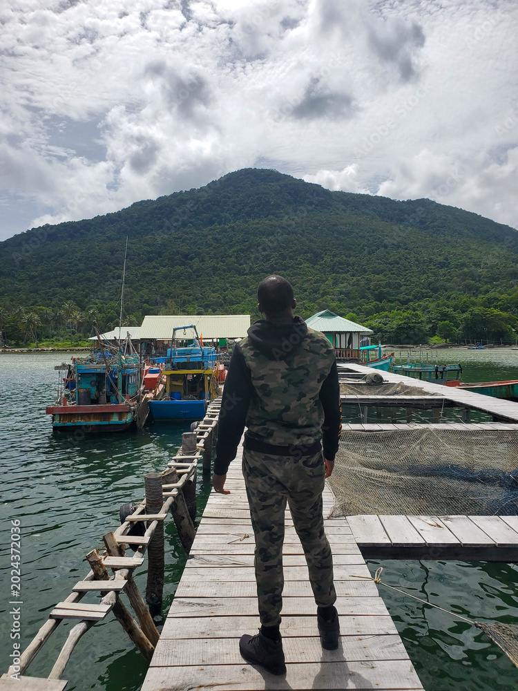 Fototapeta premium Man in camouflage standing on wooden pier overlooking water and fishing boats