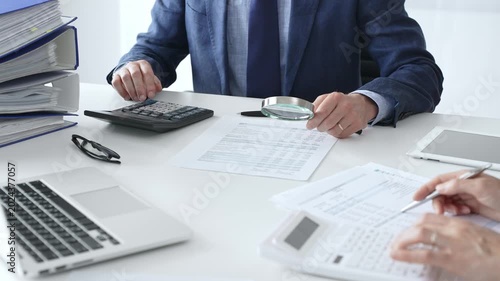 Male financial analysts examining documents, using magnifying glass and calculator while studying corporate data at workplace with a female colleague. Audit and taxes concept