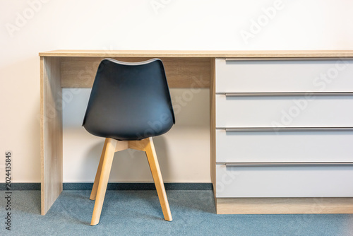 Wooden desk with dark plastic ergonomic chair, back view. Minimalistic design. Modern interior. Comfortable furniture workplace. Table with drawers.