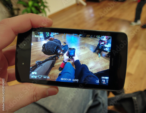 A person's hand holds a smartphone displaying a first-person shooter game scene with multiple players engaged in combat within a dimly lit interior.