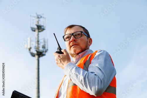 Mobile network technician conducting a field audit of cellular mast. Telecommunications engineer wearing orange safety vest using a digital tablet, talking on walkie-talkie. 5G cell phone tower.