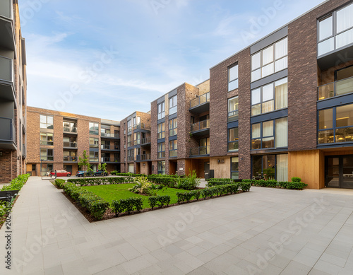 A modern residential complex features a well-maintained courtyard with lush greenery and contemporary brick architecture under a clear blue sky.