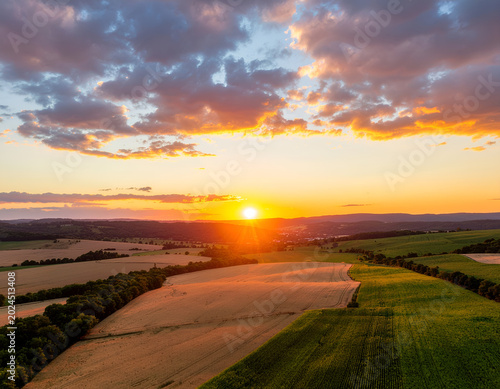 A breathtaking aerial view captures a vibrant sunset over rolling fields and lush green landscapes, creating a stunning display of natural beauty.