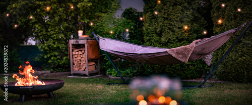 Hammock with fairy lights in garden. Cozy evening atmosphere with a campfire in a fire bowl. Background for leisure activities for outdoors.