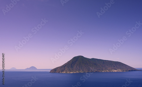 Obraz na plátně A serene view of Salina Island in the Tyrrhenian Sea, with calm turquoise waters gently lapping against a rocky shoreline under a vivid purple sky at dusk