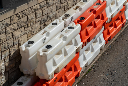 Φωτογραφία Stack of interlocking orange and white plastic water-filled traffic barriers neatly stored beside a textured stone wall on a sunny day, ready for road safety or construction work
