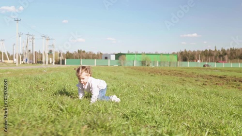 Adorable toddler girl running happily across a sunny green meadow, then stumbling and falling onto the soft grass, but she continues to smile and crawl towards the camera. Slow motion, 4K