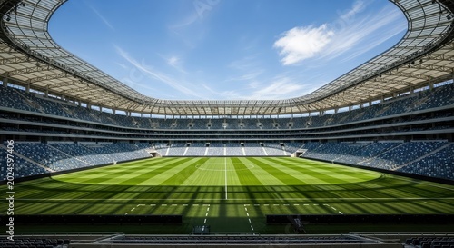 A massive contemporary arena featuring pristine green grass under a clear blue sky with intense sun lighting illuminating the empty seats, field, seats, turf
