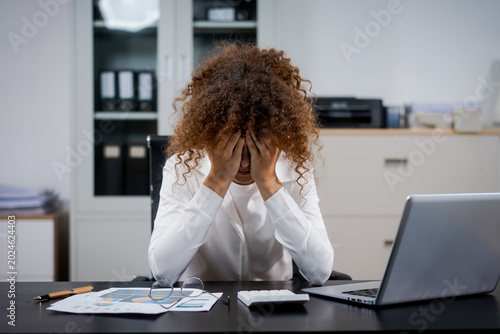 Stressed office worker with curly hair holding head at desk, laptop and paperwork in modern workspace, conveying burnout, deadlines, and financial or work pressure.