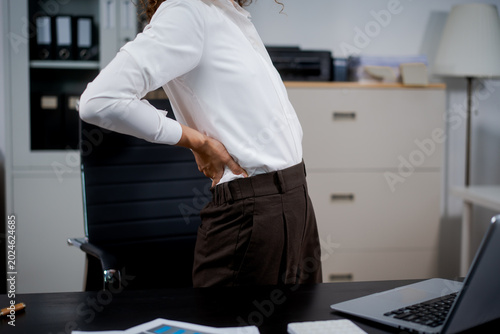 Woman in office chair stretching neck and shoulders at desk, glasses, curly hair, blurred modern workplace background; concept of neck pain relief, ergonomics, work break.