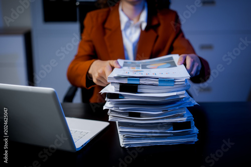 Business professional in office holding a report over a tall stack of paperwork beside a laptop, showing administrative overload, auditing, compliance, and workflow management.