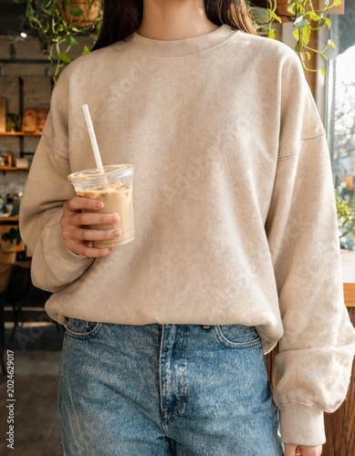 A person wearing a cream-colored sweatshirt and blue jeans, holding a plastic cup with a straw, filled with a light brown beverage, possibly iced coffee, with a hint of greenery in the background