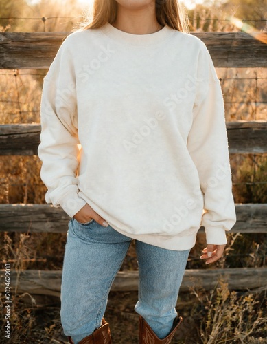 A person wearing a cream-colored sweatshirt and blue jeans standing in front of a wooden fence with warm sunlight in the background. The person's hands are in their pockets.