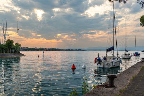 Scenic view of sailboats moored in calm waters at sunset, with a shoreline featuring trees and distant mountains in Peschiera del Garda, Italy