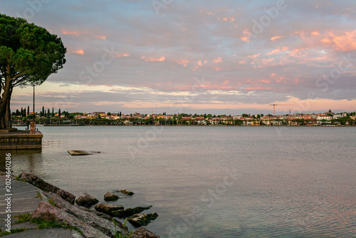 Scenic view of Lago Garda in Italy featuring calm waters, rocky shoreline, lush trees, and a picturesque town under a colorful sky during Sunrise